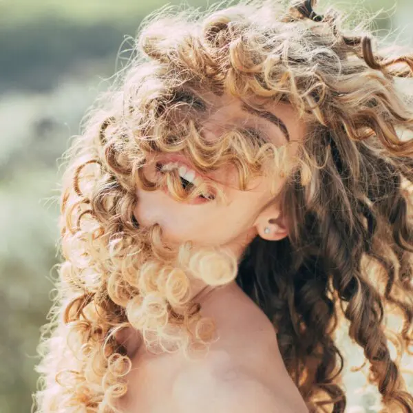 A radiant person with curly blonde hair joyfully swaying in the breeze creates a playful and lively atmosphere against a blurred green backdrop.