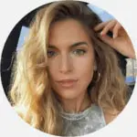 A woman with long, straight, light brown hair and light skin smiles at the camera. She is wearing a white top and stands in front of a dark background that includes a colorful wall and a framed picture. Her radiant look is courtesy of the best hair salon Cary NC has to offer.