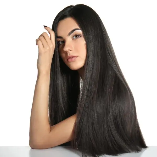 A woman with long, sleek black hair sits against a white background. She rests her chin on her hand, looking directly at the camera with a calm expression. Her glossy hair, a testament to effective hair treatments, flows over her shoulders and down the table.