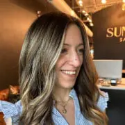 A woman with long, wavy brown hair smiles inside a salon. She is wearing a blue and white striped blouse. The salon has a modern design with warm lighting and a visible sign in the background.