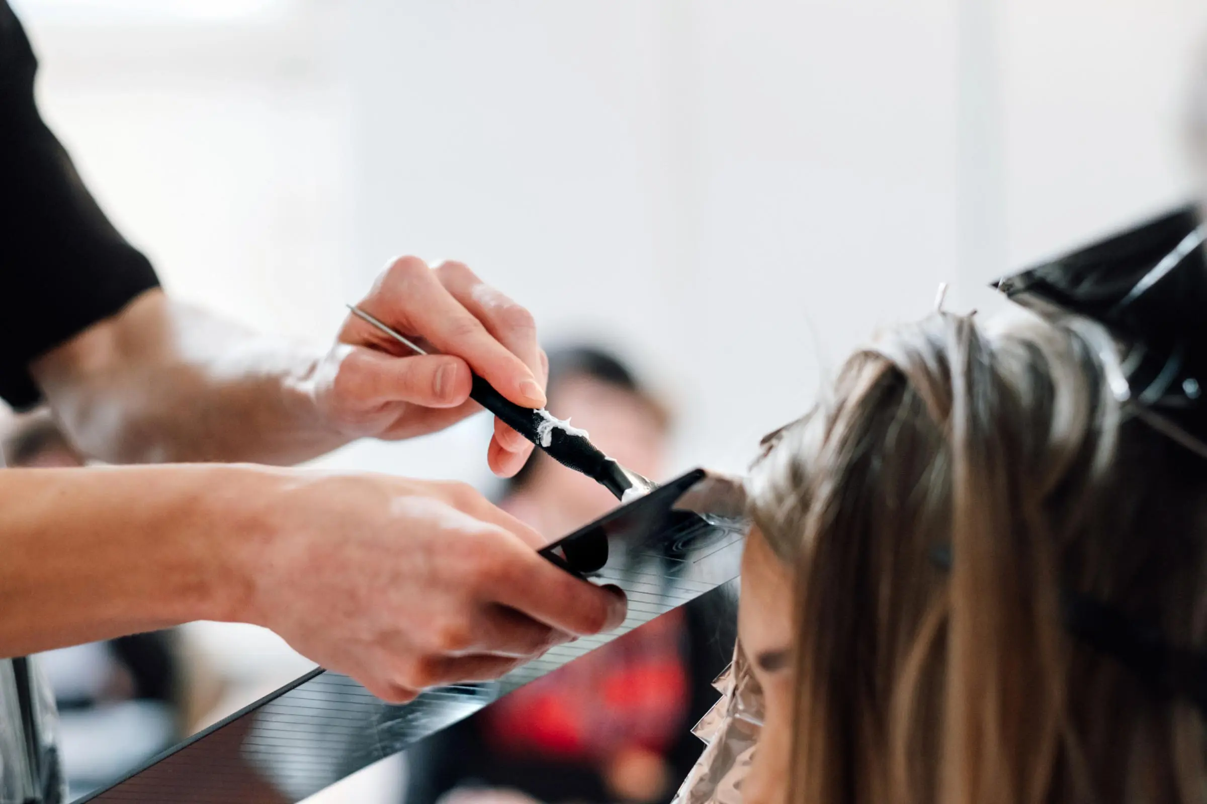 A hair color specialist skillfully applies dye to a client's hair using a brush and tray. The client is seated with foils in her hair, showcasing the professional coloring process. The softly blurred background ensures all focus remains on the expert hands and their meticulous work.