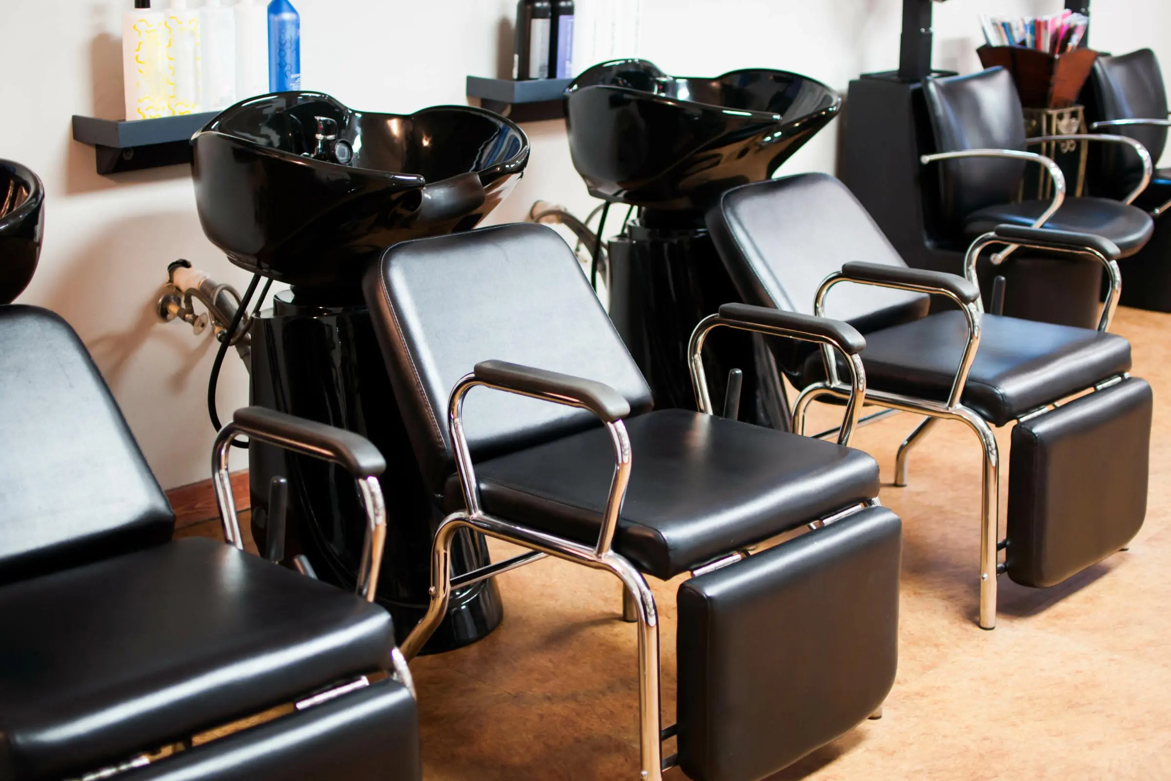 A row of modern black salon chairs with footrests in front of washbasins creates a sleek, inviting path to cosmetology excellence. The clean and organized setup features shelves brimming with hair products, echoing the dedication needed to earn a cosmetology license.