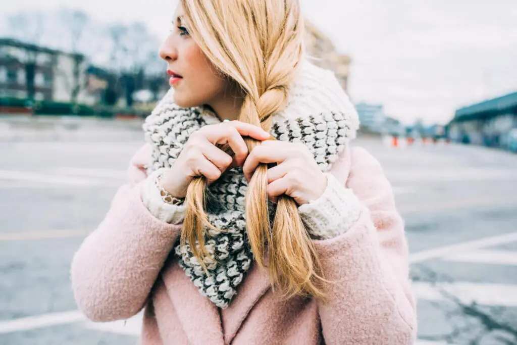 A woman wearing a pink coat and knitted scarf braids her long blonde hair, showcasing one of many easy hairstyles. She stands outdoors on a cloudy day, with blurred urban surroundings in the background.