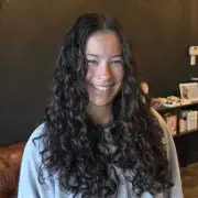 A person with long, curly dark hair smiles while standing indoors. They are wearing a light-colored shirt, and the background features a dark wall with shelves holding various items.