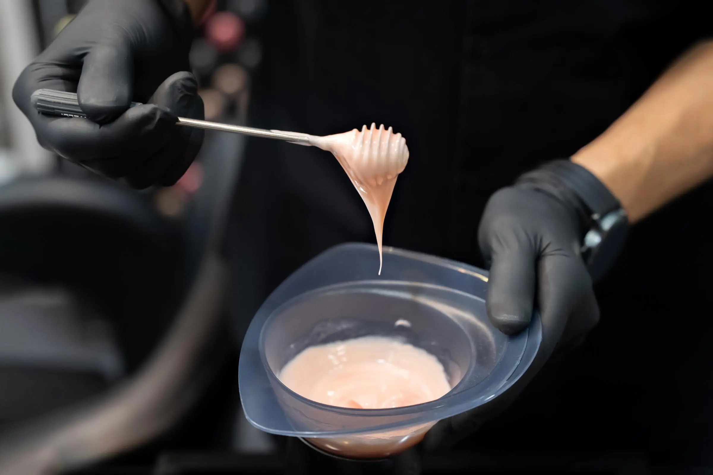 A colorist expertly wearing black gloves holds a brush coated in pastel pink hair dye, poised above a small bowl brimming with the vibrant hue.
