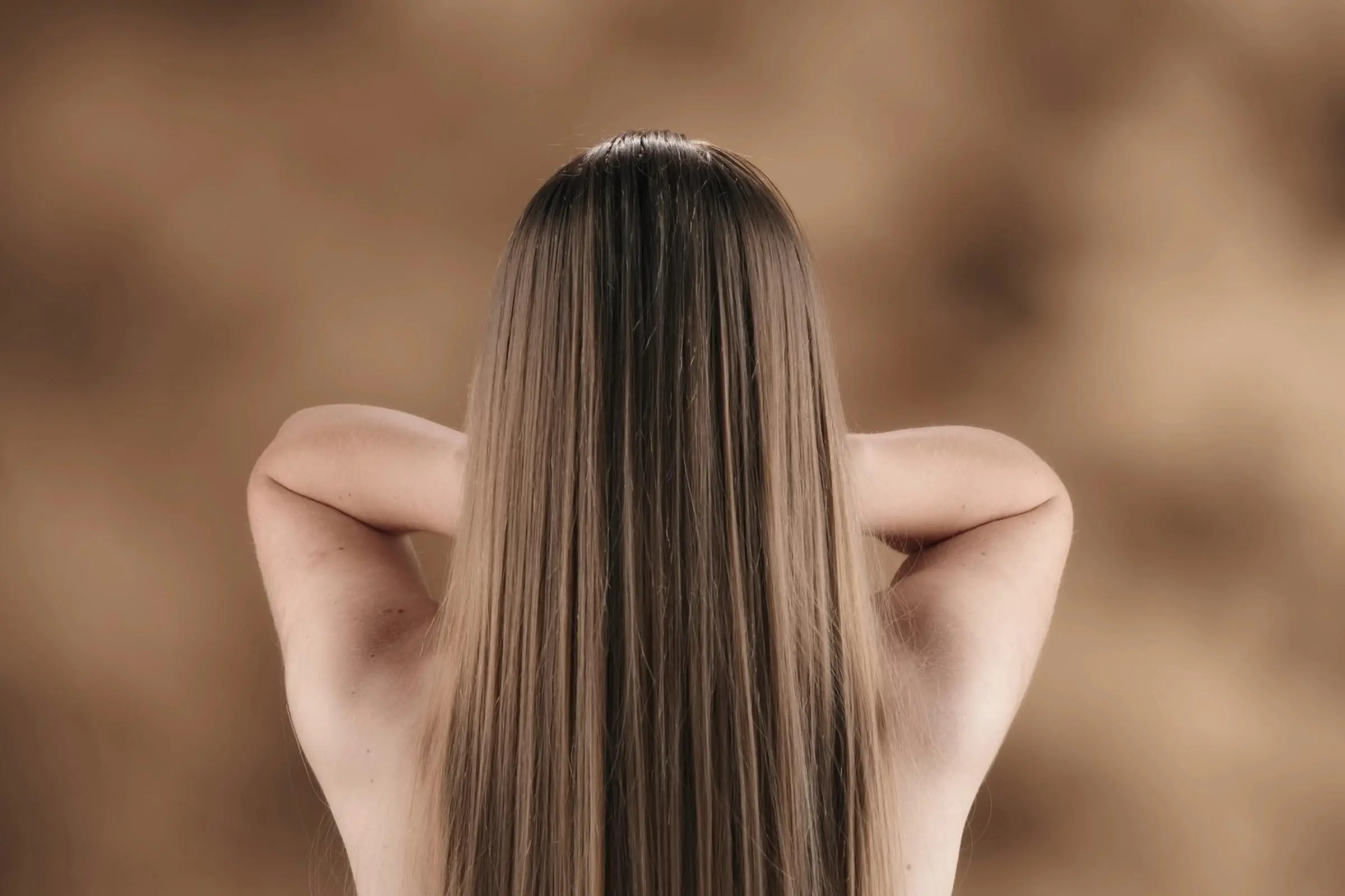 A person with long, straight, dirty blonde hair stands with their back to the camera, arms raised and hands resting lightly on the back of their neck against a soft, brown blurred background.