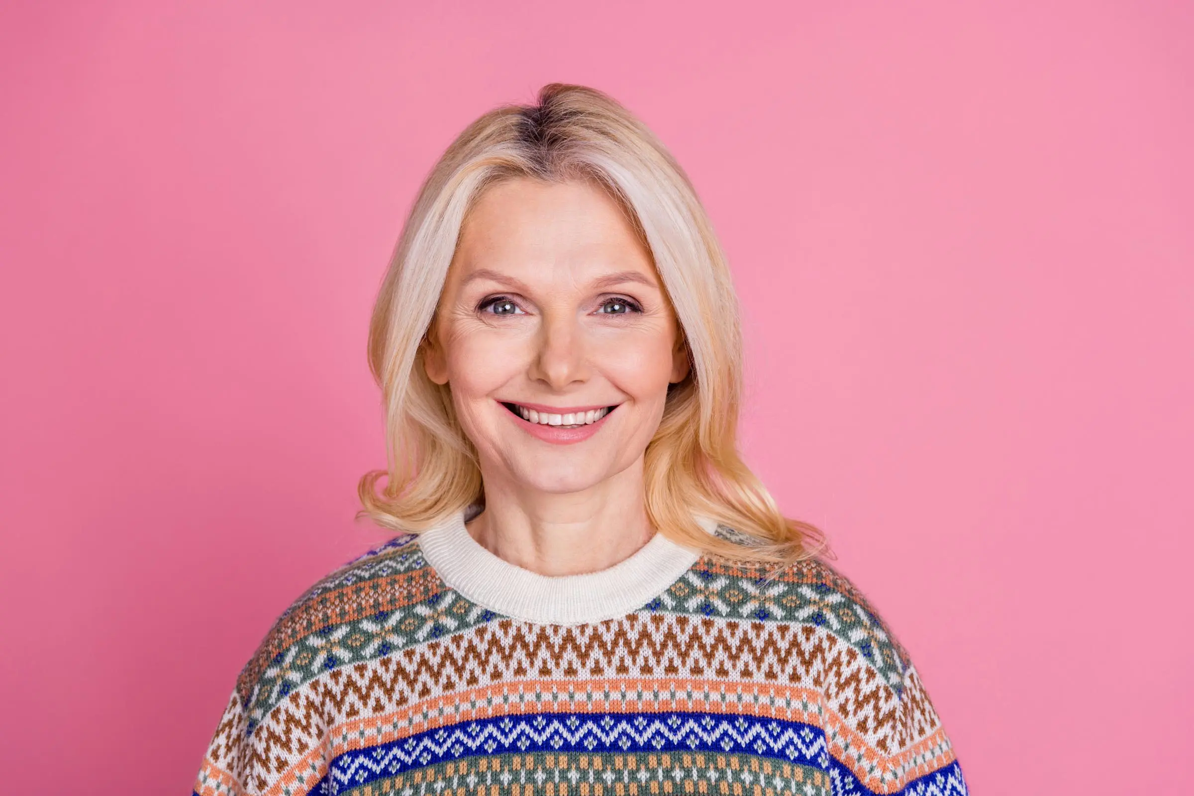 A smiling woman with long blonde hair, showcasing her stylish patterned sweater, stands gracefully against a pink background, leaving one to wonder: how long does grey coverage last for maintaining such vibrant locks?