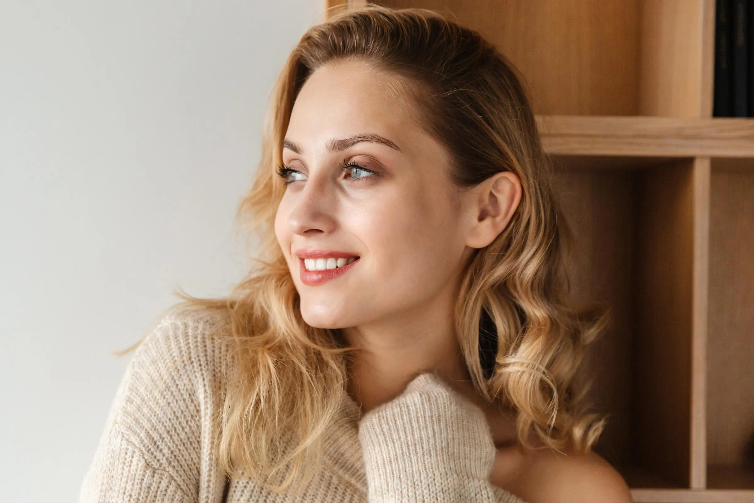 A woman with wavy, blonde hair smiles softly, looking to the side. Her cozy, light-colored knit sweater complements her look, while subtle blonde hair touch-ups add a fresh feel as she stands in front of a wooden bookshelf, crafting a warm and relaxed atmosphere.