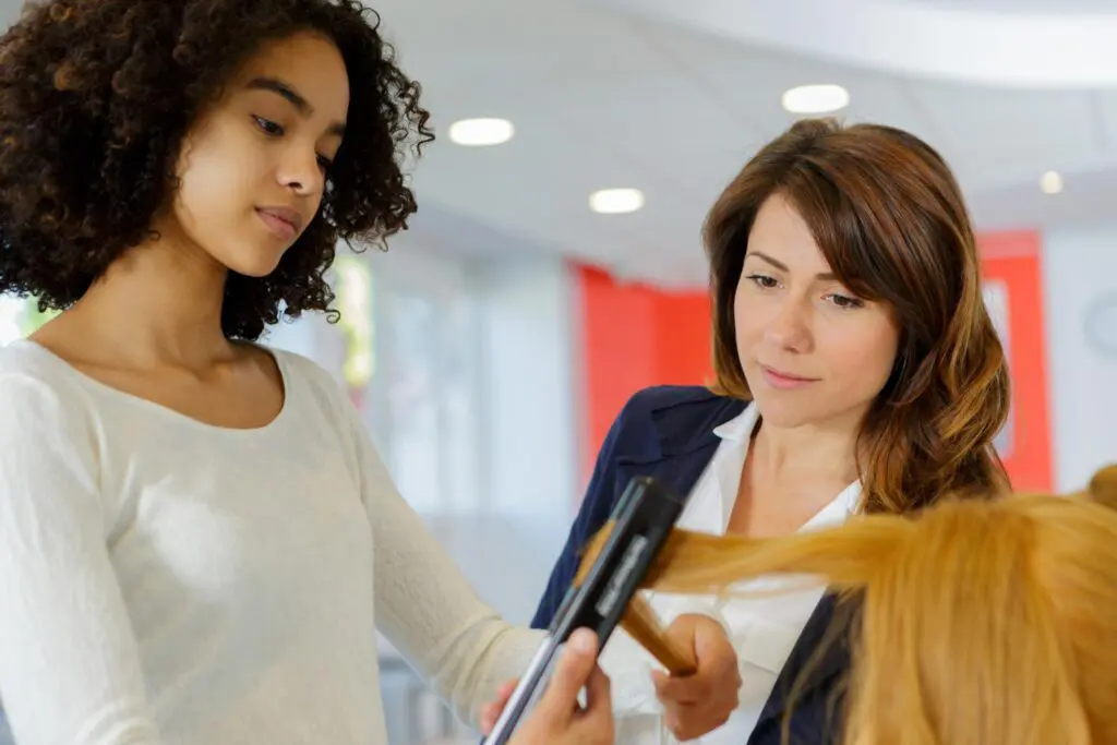 A skilled salon apprentice with curly hair uses a hair straightener on another person's locks, while a woman with straight hair observes intently. The bright lighting and red accents create the perfect setting to enhance your visit.