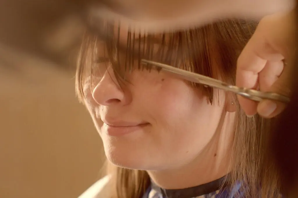 A person with long brown hair is getting a precision haircut. Scissors are expertly trimming their bangs while they sit, smiling slightly, covered in a blue and white cape. Their eyes are obscured by hair being cut—a perfect snapshot of modern lifestyles embracing stylish solutions.