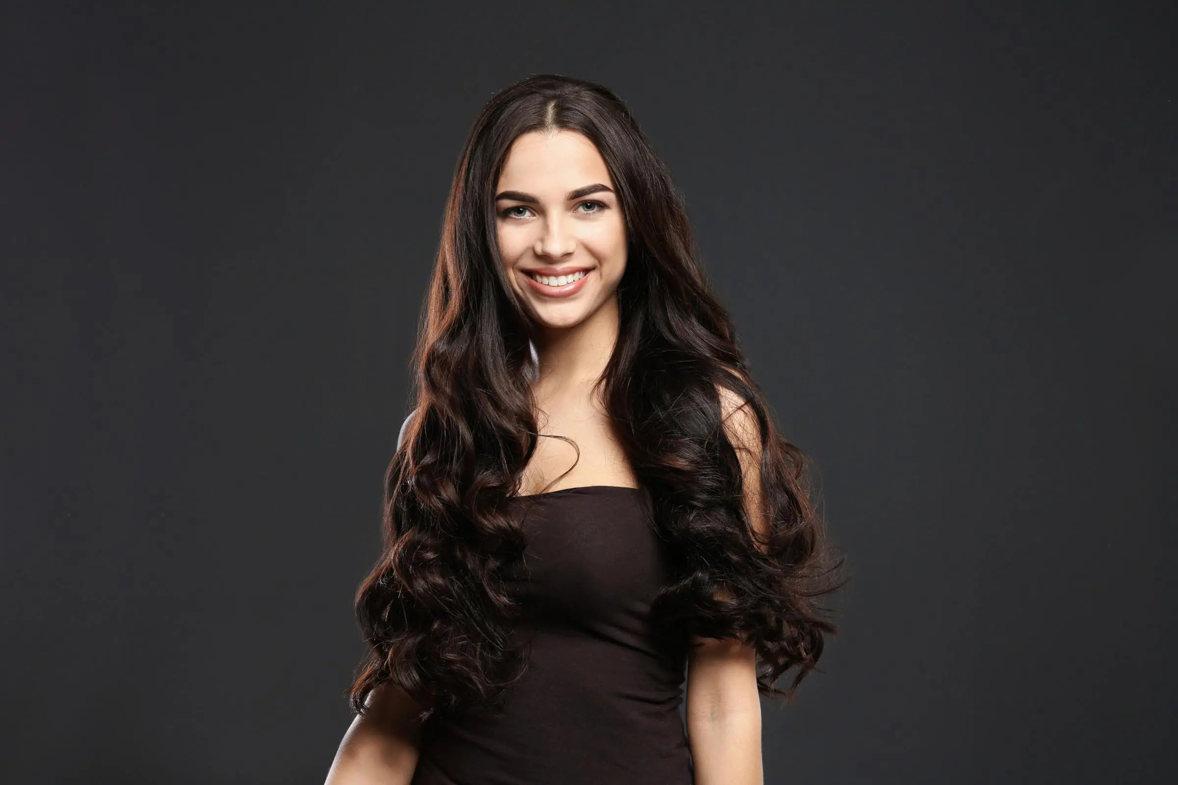 Smiling woman with long, wavy dark hair wears a sleeveless black top, standing against a dark background.