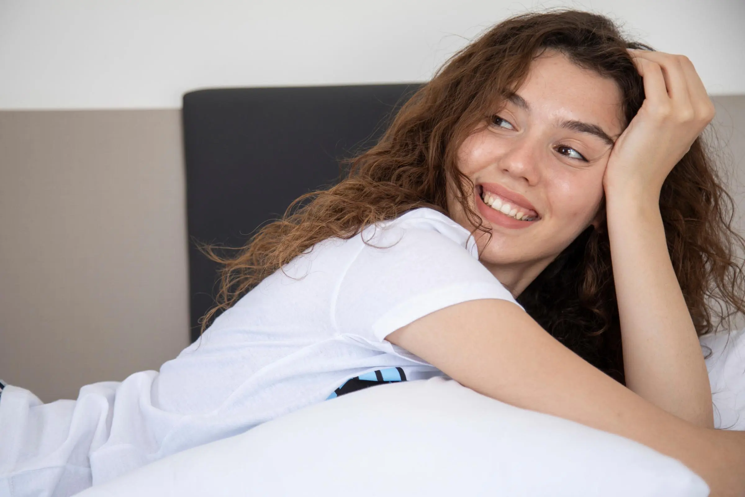 A person with long curly hair lies on a bed, smiling and looking to the side. They are wearing a white t-shirt. The setting appears to be indoors with soft lighting, showcasing how to sleep comfortably with curly hair while maintaining those luscious curls.