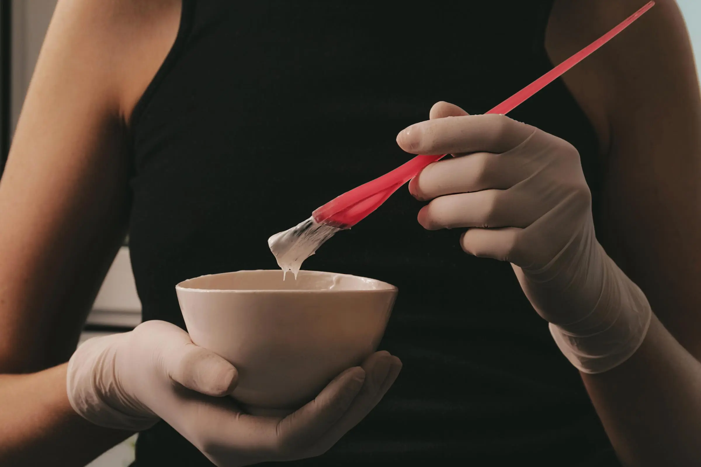 A Hair Colorist wearing a black shirt and white gloves holds a small, light-colored bowl and a red brush with white paint on the bristles, ready to share expert hair care tips. The setting appears to be indoors.