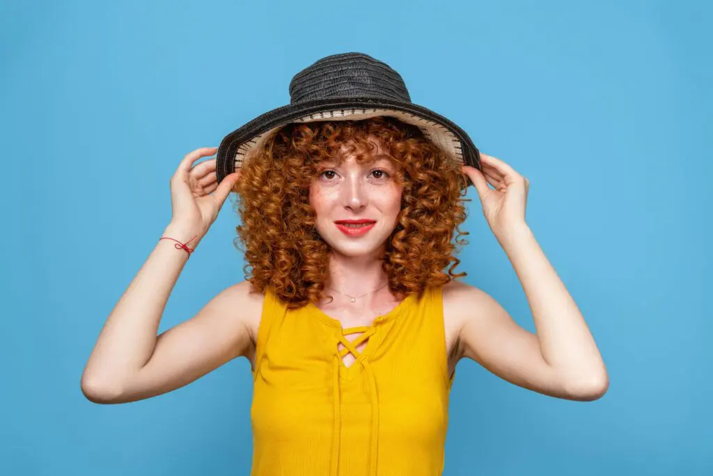8 Tips for Maintaining Healthy Permed Hair_edited A person with curly red hair, reminiscent of wild perms, smiles while holding the brim of a wide-brimmed black hat. They are wearing a yellow sleeveless top and are set against a plain blue background.