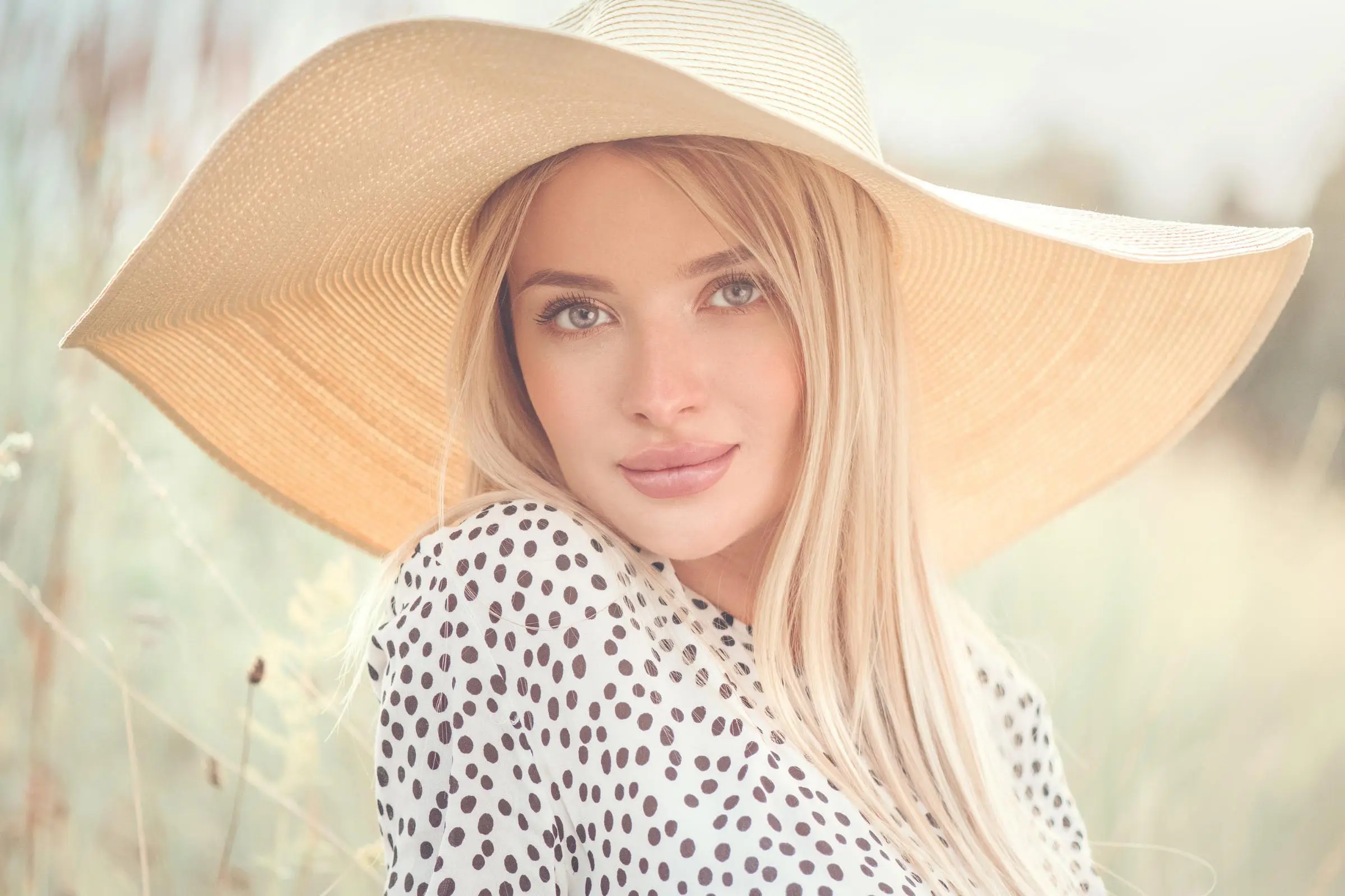 A woman with long blonde hair is wearing a large, wide-brimmed straw hat and a white blouse with black polka dots. She is outdoors, surrounded by soft-focus greenery, giving the image a warm, serene feel.