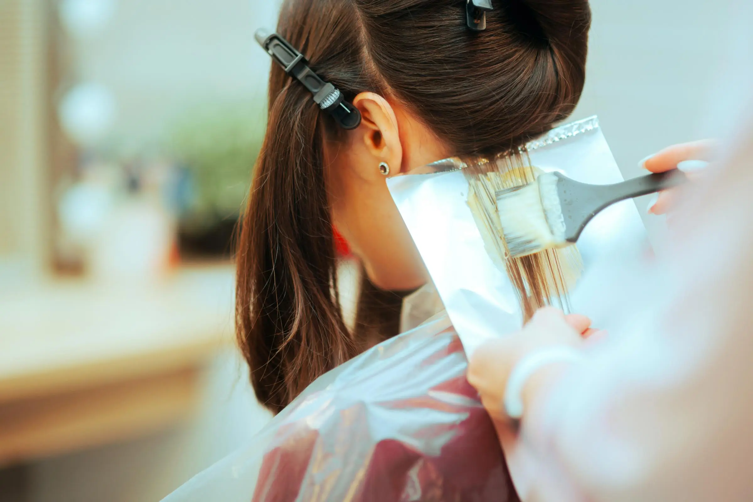 A person sits in a salon chair, immersed in a color technique as sections of hair are clipped away. A stylist deftly applies dye using a brush and foil, with the background softly blurred to highlight the precision of the hair treatment process.