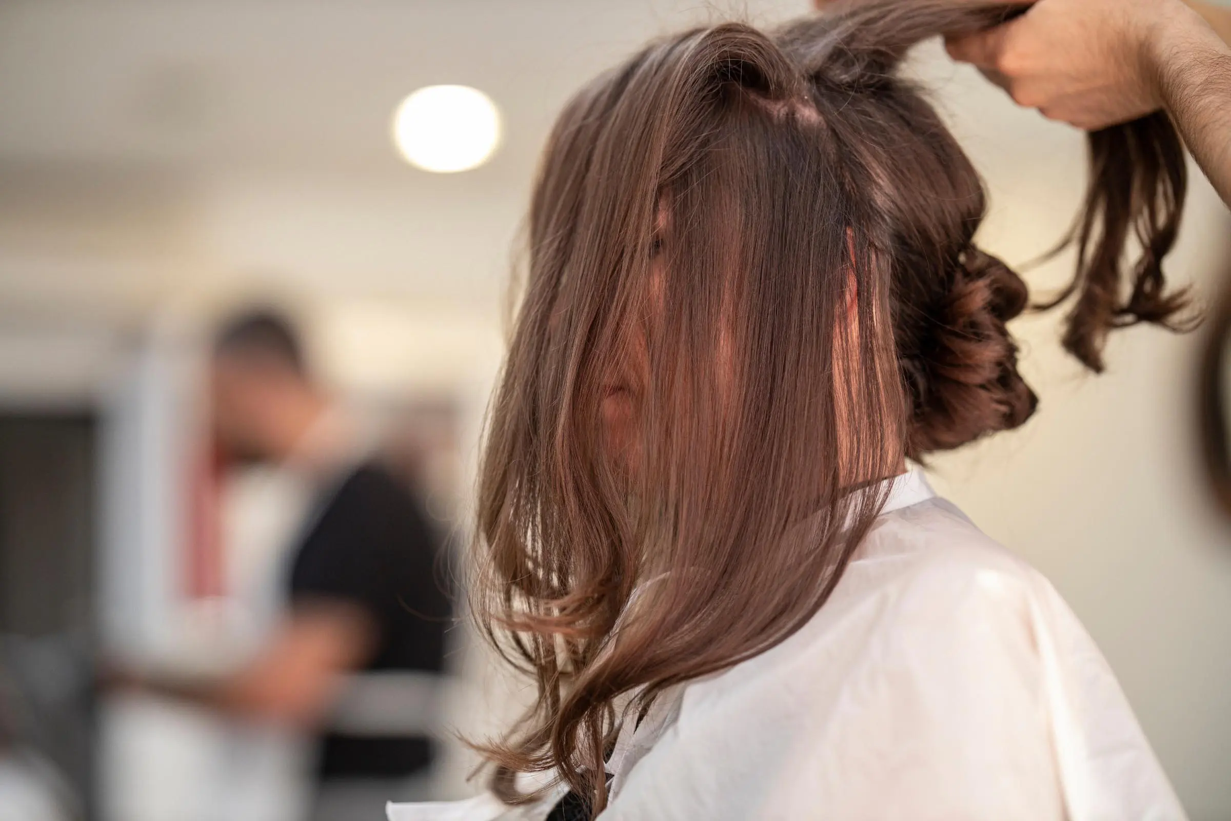 Person with long brown hair, partially covering their face, is sitting in a salon chair with a white cape. A stylist, using salon tricks, is pulling up sections of their hair for an amazing blowout. The background is softly blurred with another person standing and working.
