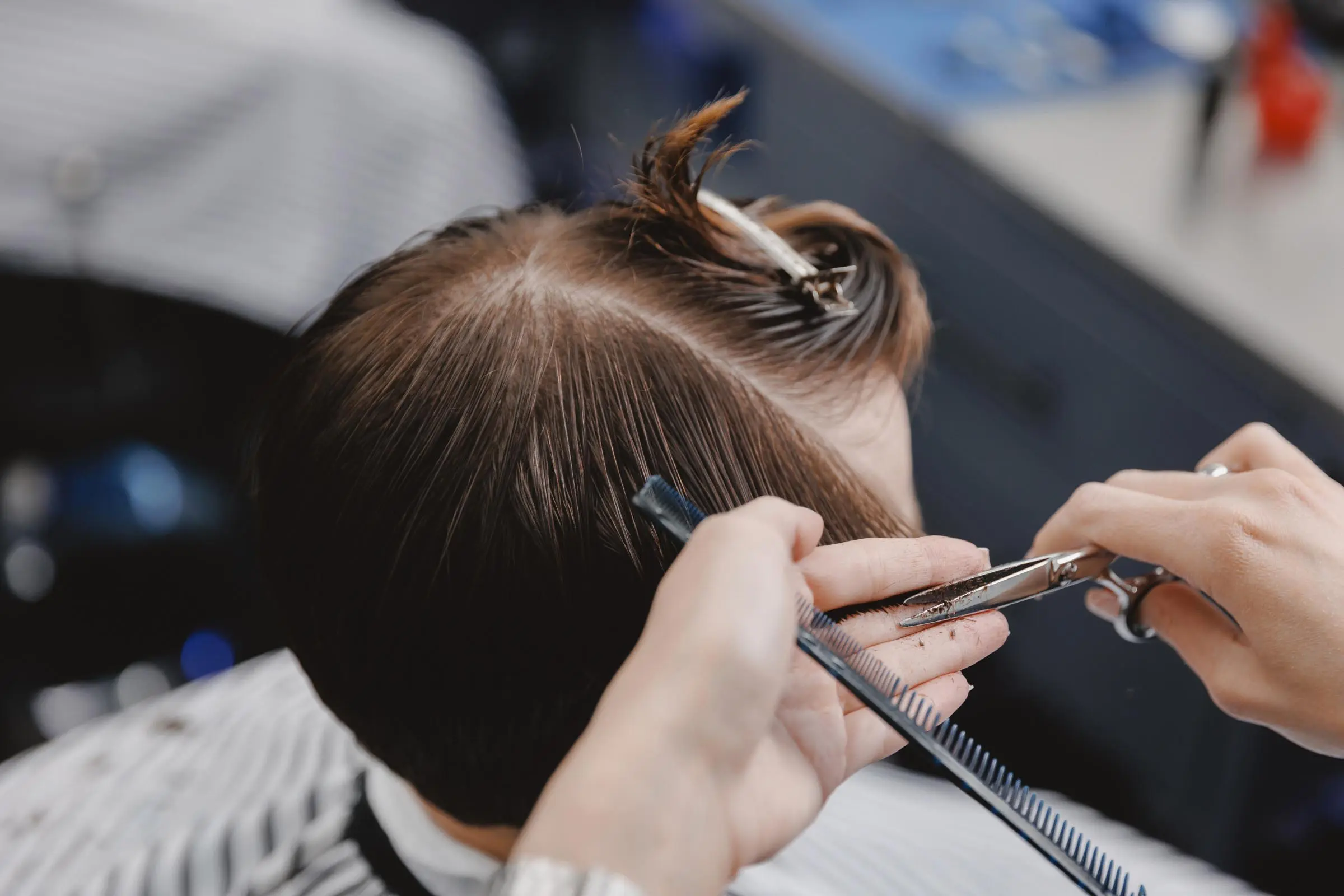 A hairdresser, chosen with care, uses scissors and a comb to trim a person's brown hair in a salon, with hair sectioned and clipped on top—highlighting the importance of choosing a hair stylist you trust.