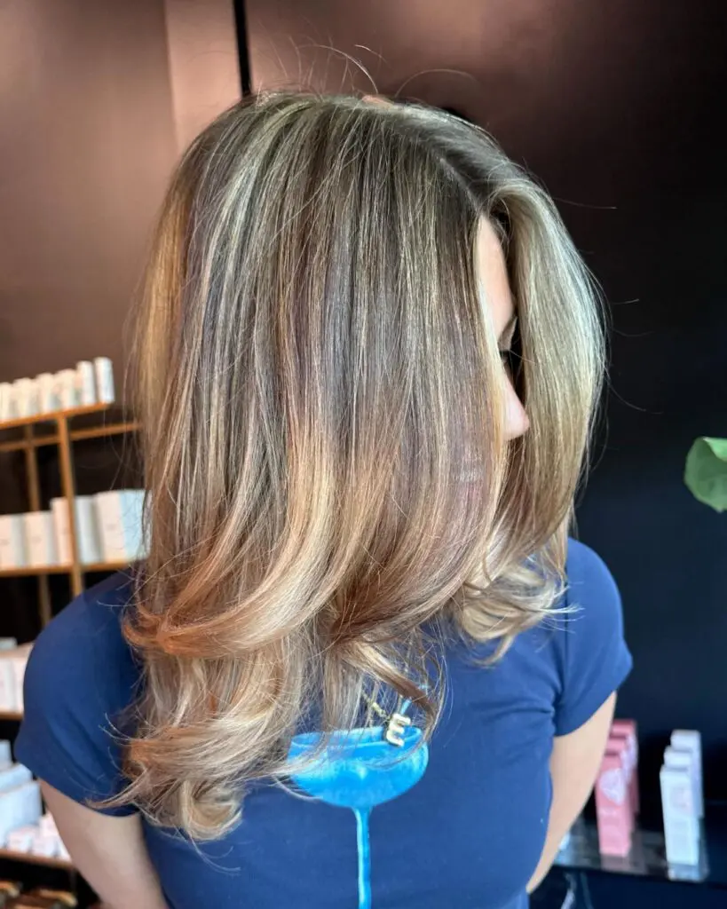 Dimensional Honey Blonde Highlights with a Bouncy Layered Blowout Mid-length brown hair with dimensional honey and ash blonde highlights, styled in a sleek, bouncy layered blowout with flipped ends at Sunday Salon in Raleigh.