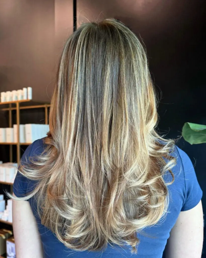 Golden Honey Highlights with Voluminous Bouncy Layers Back view of a client at Sunday Salon in Raleigh with long, heavily layered hair styled in a bouncy blowout, featuring dimensional golden honey blonde highlights and a darker natural base.