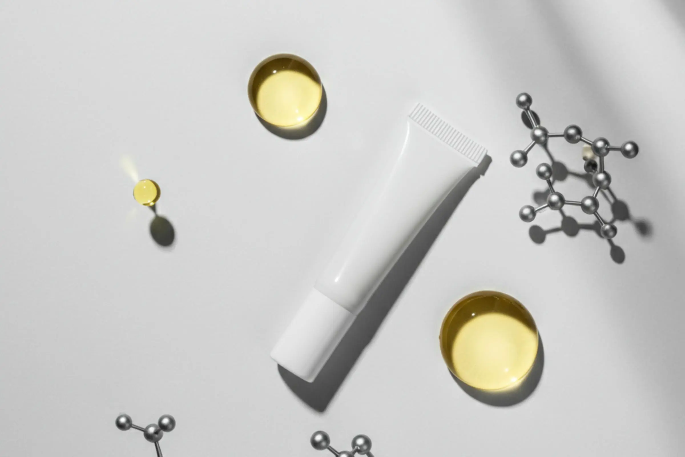 A white tube of The Ordinary Squalane + Amino Acids Hydrating Lip Balm is surrounded by small pools of yellow liquid and silver molecular structures on a light gray background.