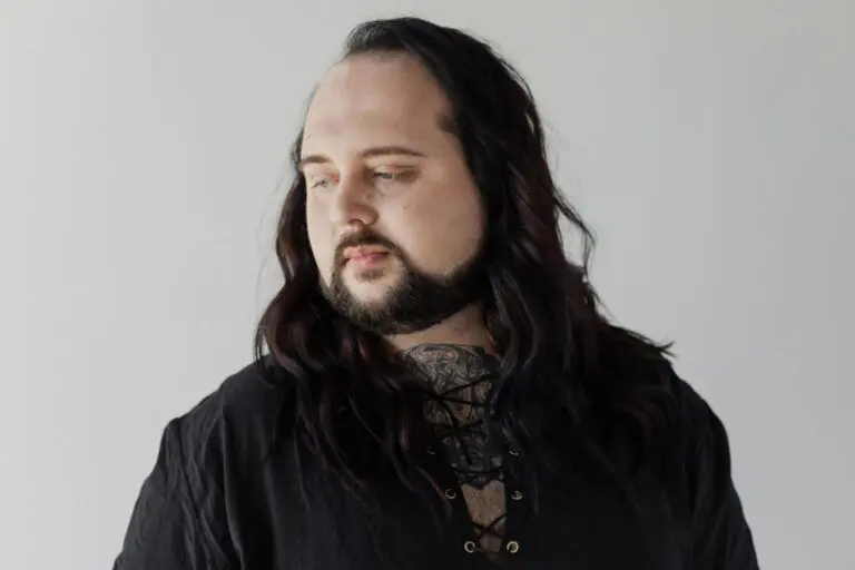 A person with long dark hair—perhaps shaped by Advanced Hair Stylist Education—a beard, and a visible chest tattoo, wearing a black lace-up shirt, looks thoughtfully to the side against a plain light background.
