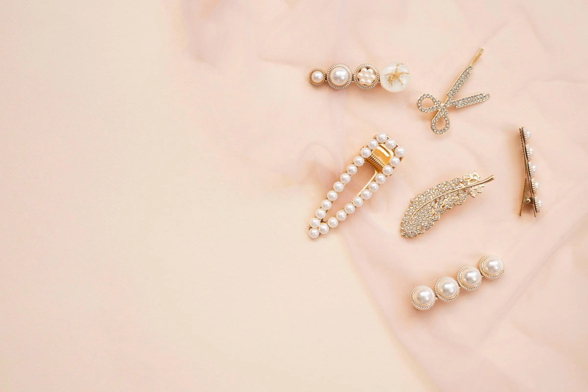 Assorted pearl and gold hair clips are arranged on a soft, light beige fabric background, showcasing elegant and decorative designs in various shapes including circles, a feather, and a bow.