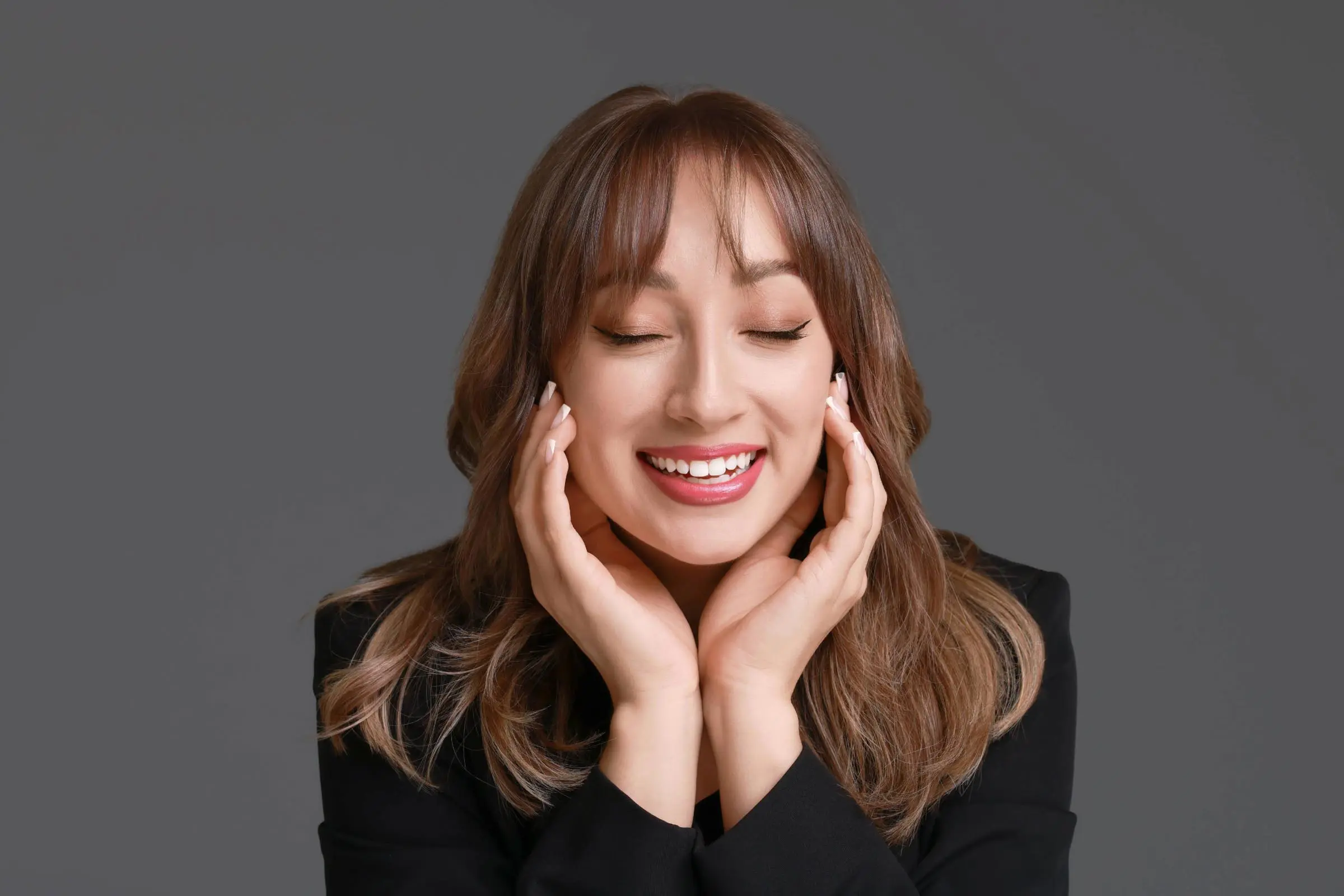 butterfly.cut A young woman with long brown hair styled in a butterfly haircut smiles with her eyes closed, gently resting her face in her hands. She is wearing a black top and is posed against a plain gray background.