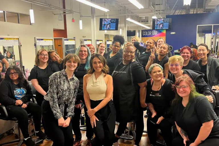 A group of smiling people, mostly women, pose together inside Sunday Salon & Aveda Chapel Hill, some wearing black salon uniforms. Mirrors, salon chairs, and a "Color Bar" sign are visible in the brightly lit background.