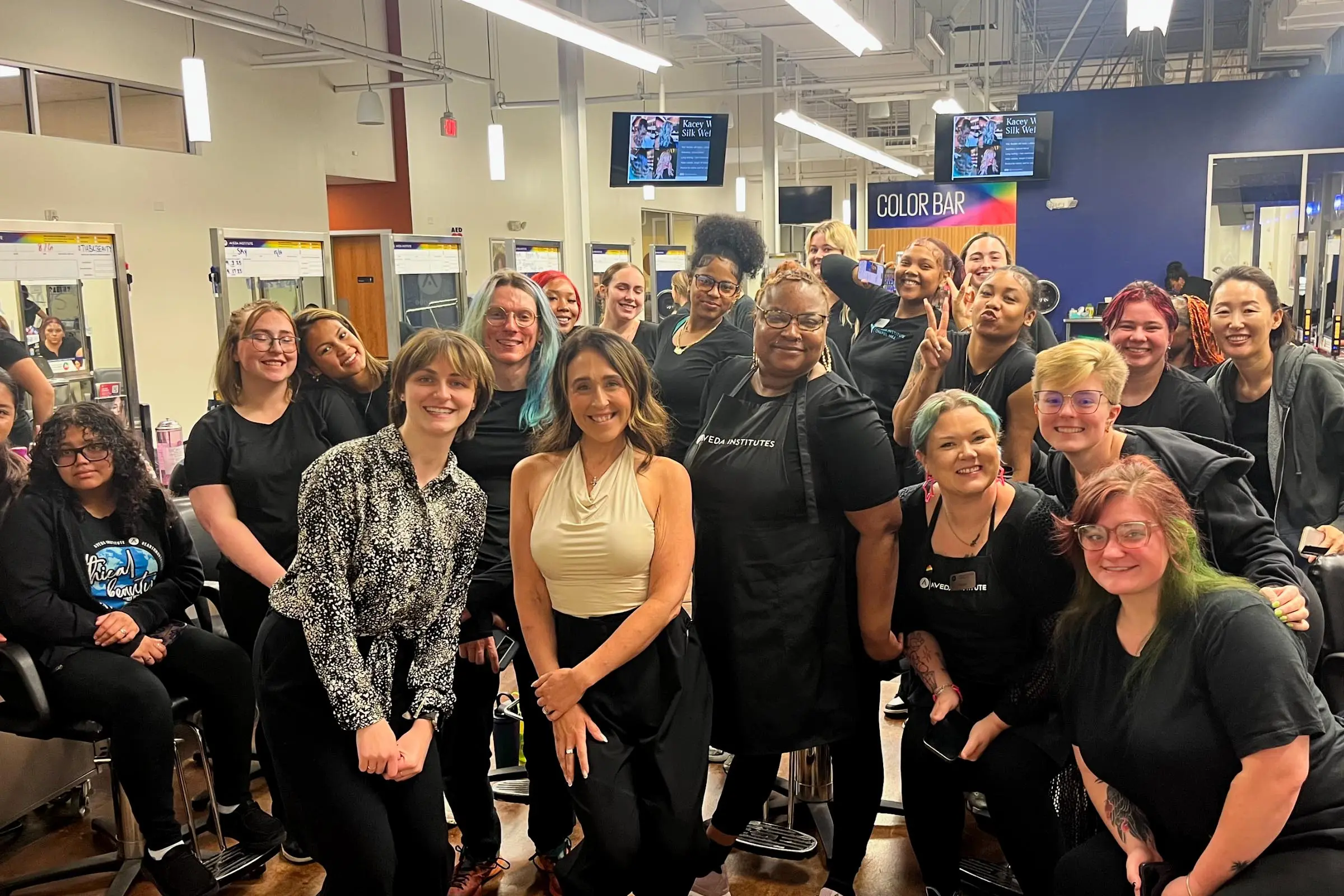 A group of smiling people, mostly women, pose together inside Sunday Salon & Aveda Chapel Hill, some wearing black salon uniforms. Mirrors, salon chairs, and a "Color Bar" sign are visible in the brightly lit background.