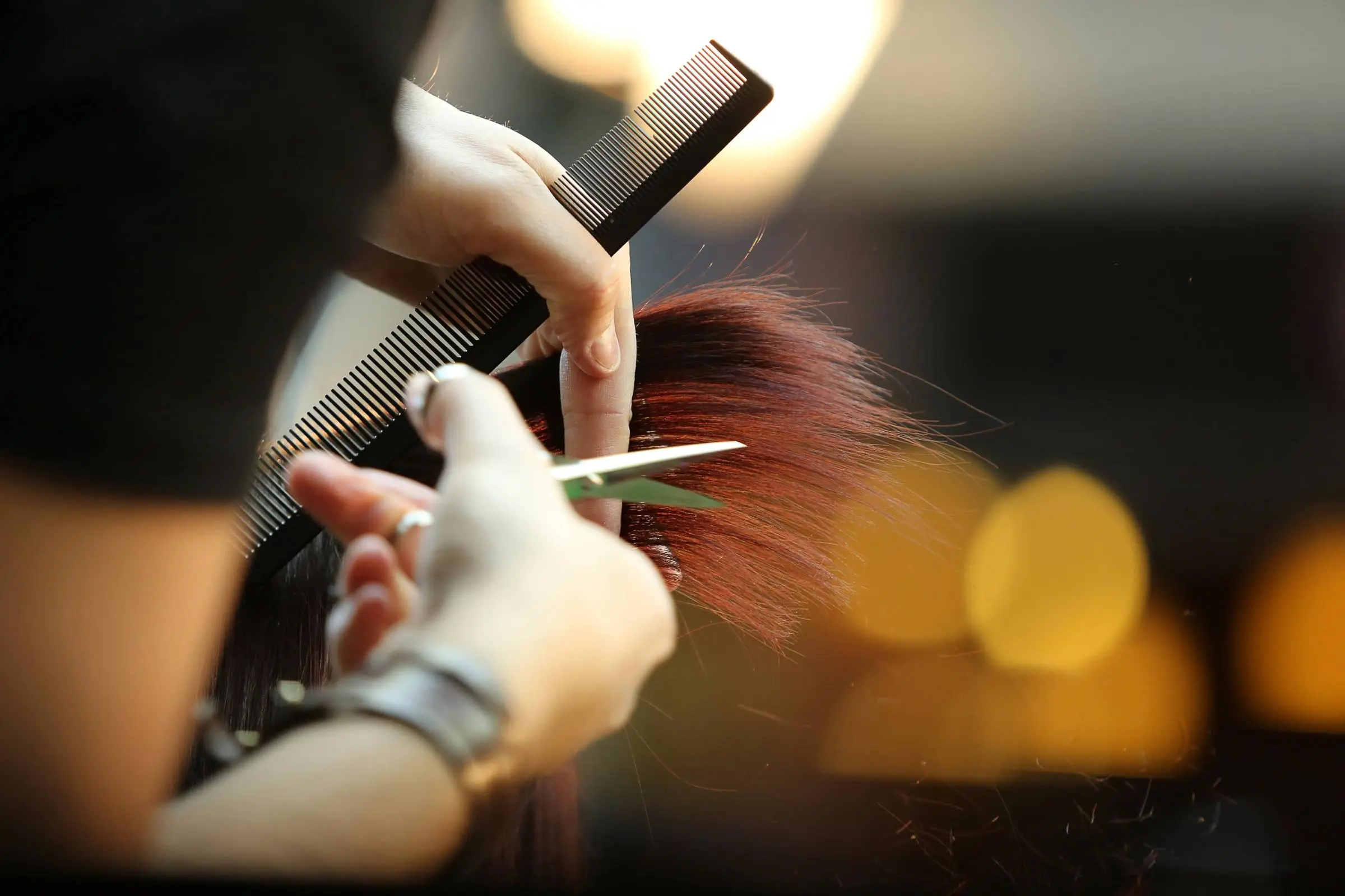 A close-up of a hairstylist’s hands cutting brown hair with scissors while holding a comb, with blurred lights in the background—perfect for those seeking haircut tips or inspiration to maintain a haircut at home.
