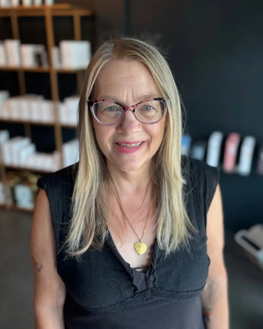 Emily Genthe-1 A smiling woman with long blonde hair and glasses stands indoors, wearing a black top and a gold heart necklace. Shelves with white and pastel items are visible in the background.