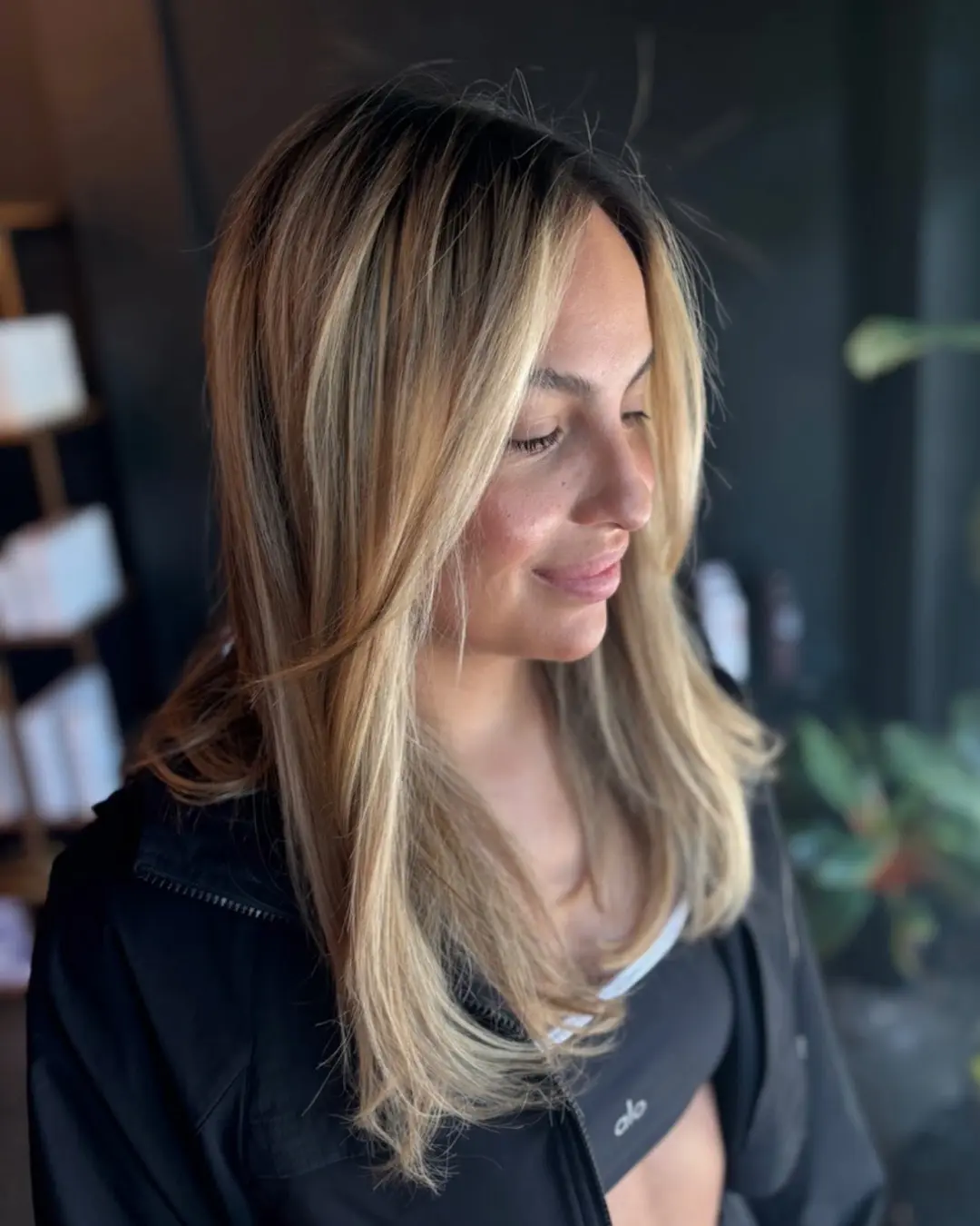 Emily Genthe-2 A woman with long, straight, blonde-highlighted hair smiles softly while looking down. She is wearing a black jacket over a black top, with blurred indoor plants and shelves in the background.