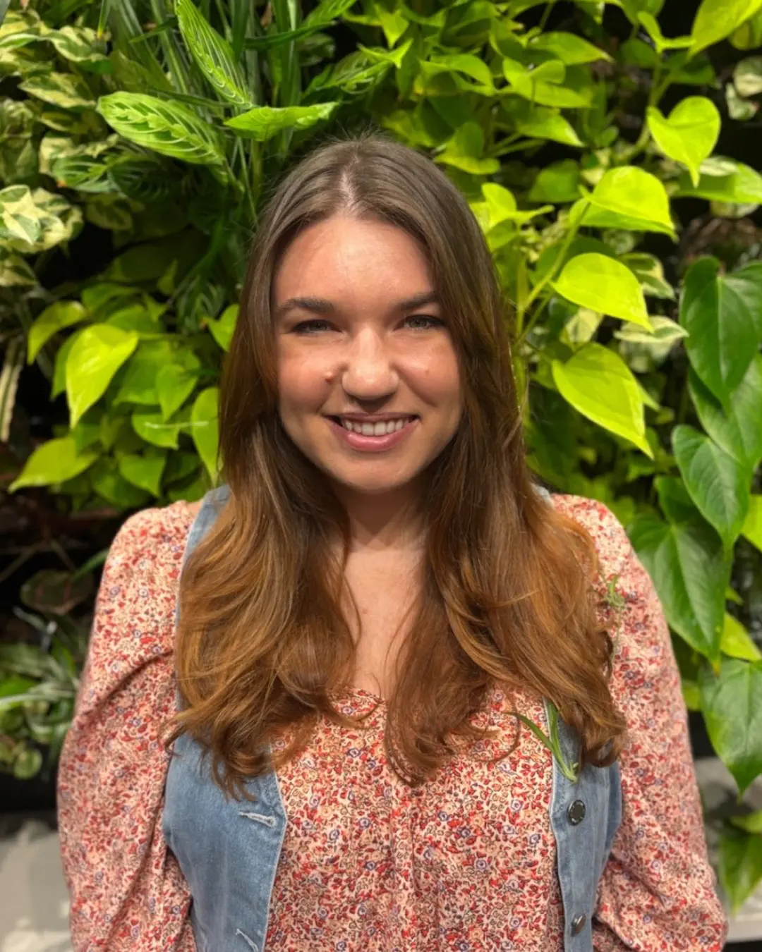 Emily Genthe-4 A woman with long brown hair, wearing a floral blouse and a blue vest, smiles at the camera while standing in front of a lush green wall of assorted plants.