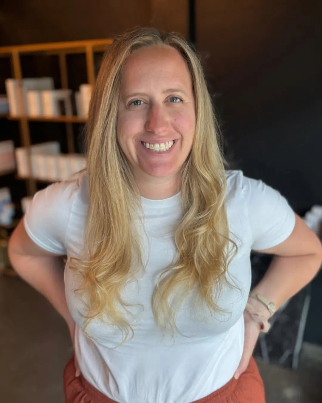 Emily Genthe-6 A person with long blonde hair, wearing a white t-shirt and rust-colored pants, smiles while standing indoors with hands on hips. Shelves and a dark wall are visible in the background.