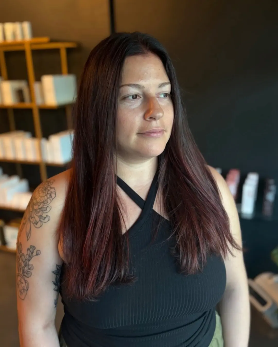 Emily Genthe-7 A woman with long, straight dark hair and a black sleeveless top stands indoors. She has a floral tattoo on her upper arm and is looking slightly to the side. Shelves with white boxes are visible in the background.