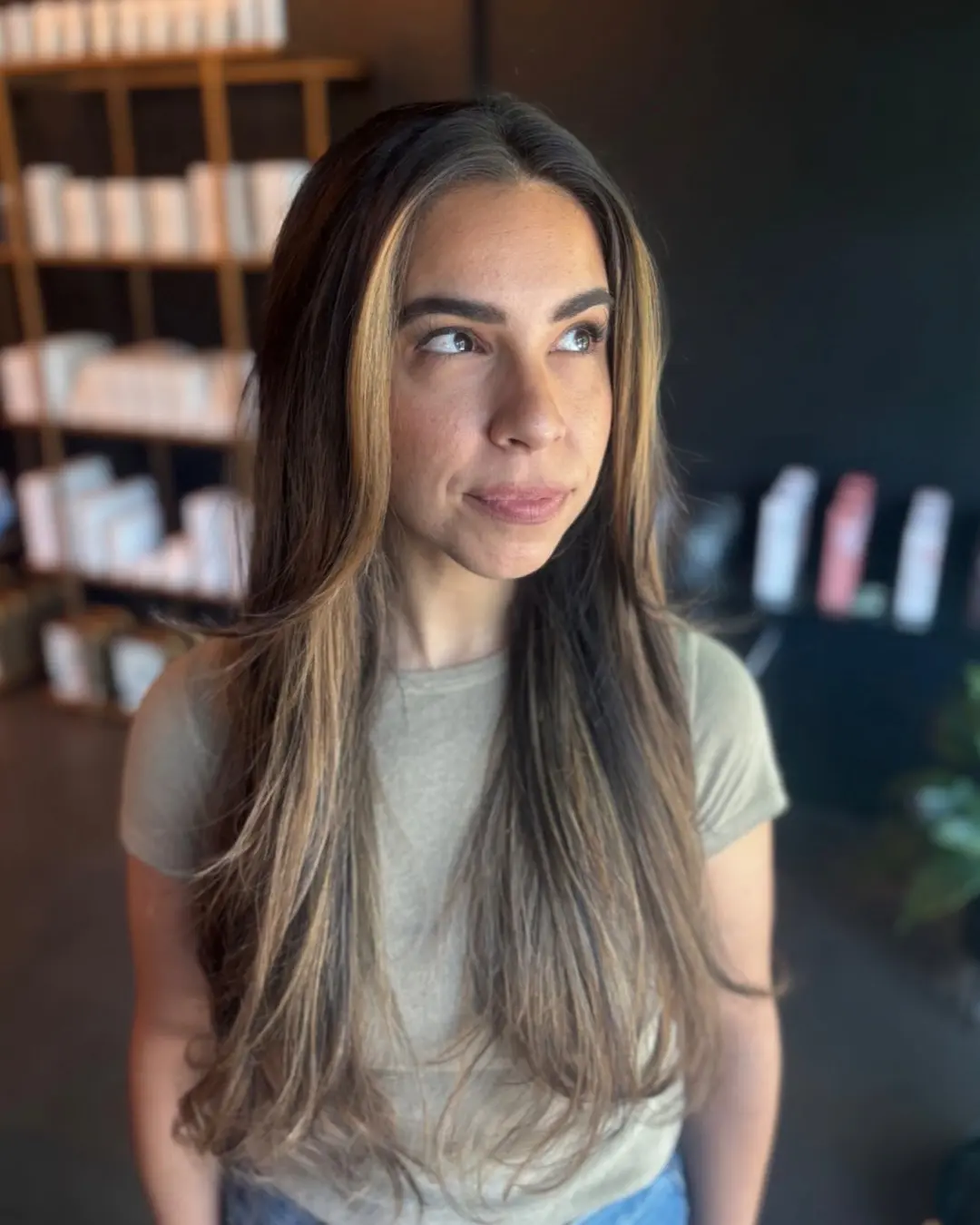 Emily Genthe-8 A woman with long, straight brown hair and blonde highlights stands indoors, looking slightly upward and to her left. She is wearing a light green short-sleeve top, with shelves and products blurred in the background.