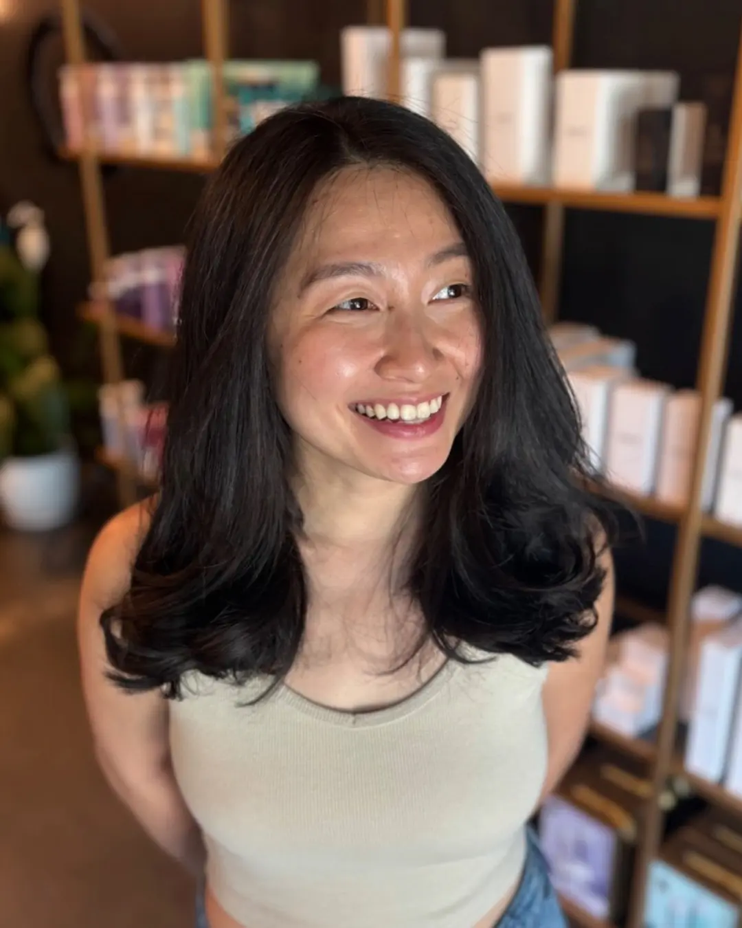 Emily Genthe-9 A woman with long, dark hair and a sleeveless beige top smiles while standing in a store with shelves of beauty or skincare products in the background.