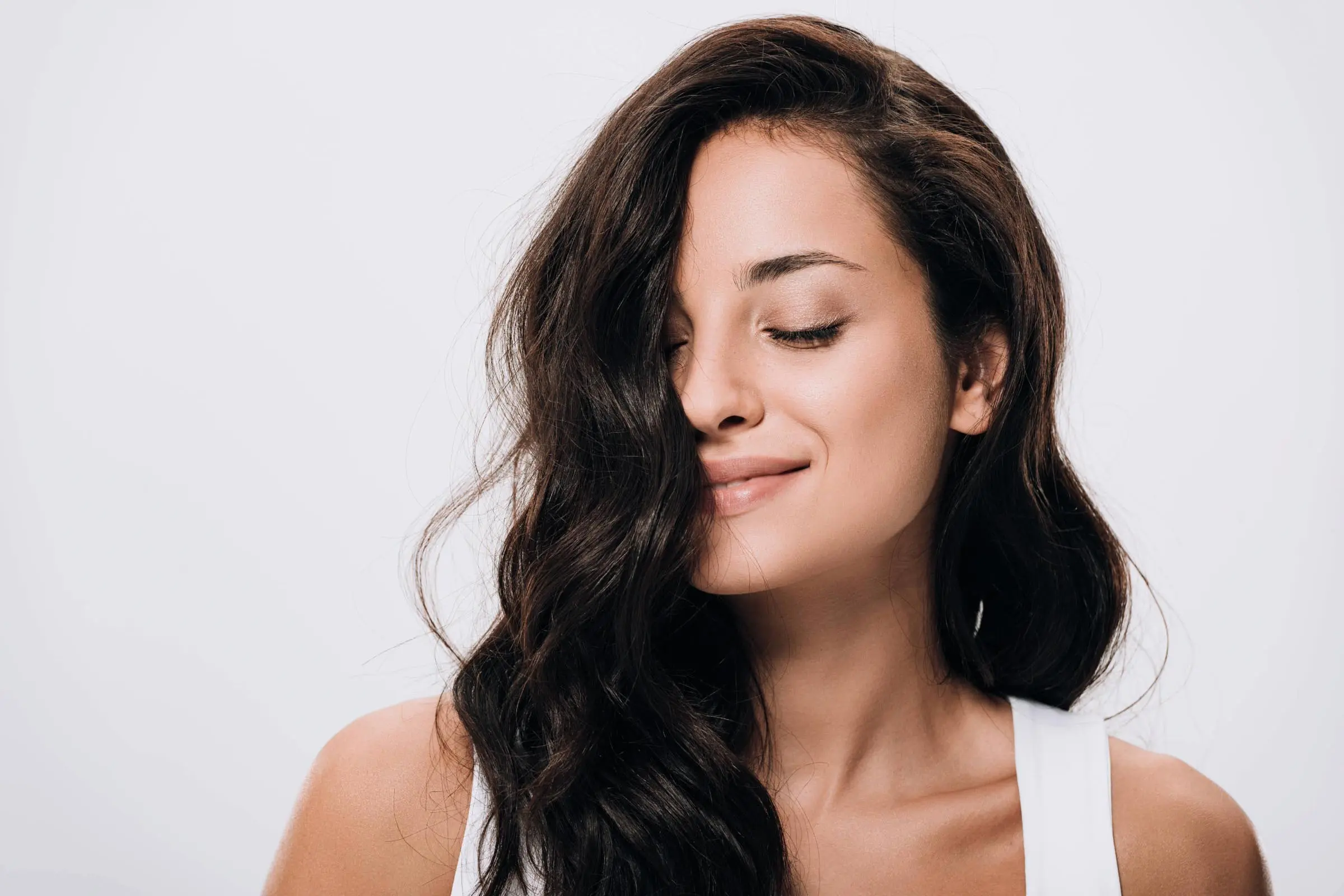 A woman with long, wavy dark hair and a slight smile poses with her eyes closed against a plain light background. She is wearing a white top.