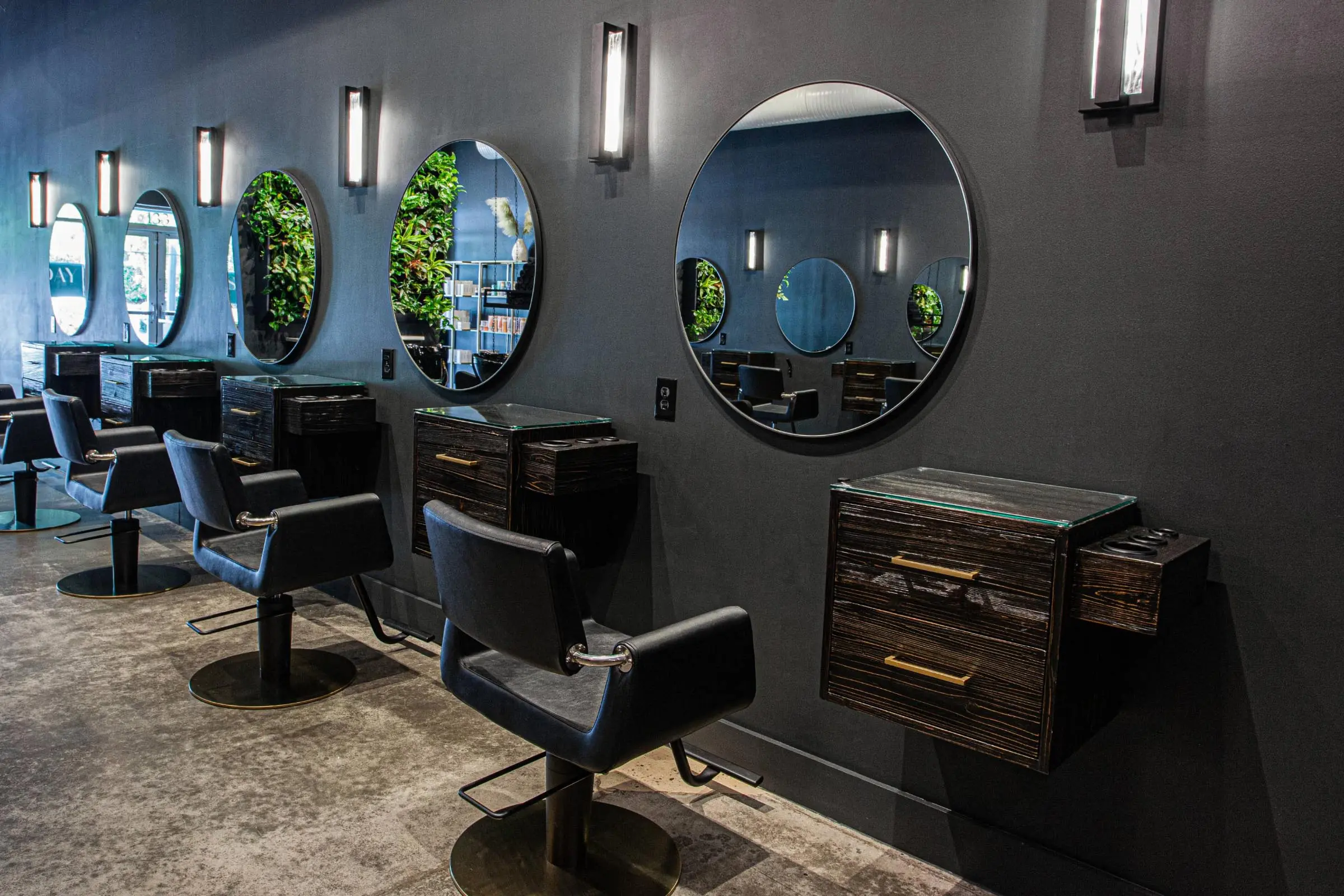 A modern hair salon offering quality salon services, with black styling chairs facing round mirrors on a dark wall. Each station features a drawer unit beneath the mirror, and lush plants are reflected. The concrete floor adds to the contemporary vibe.