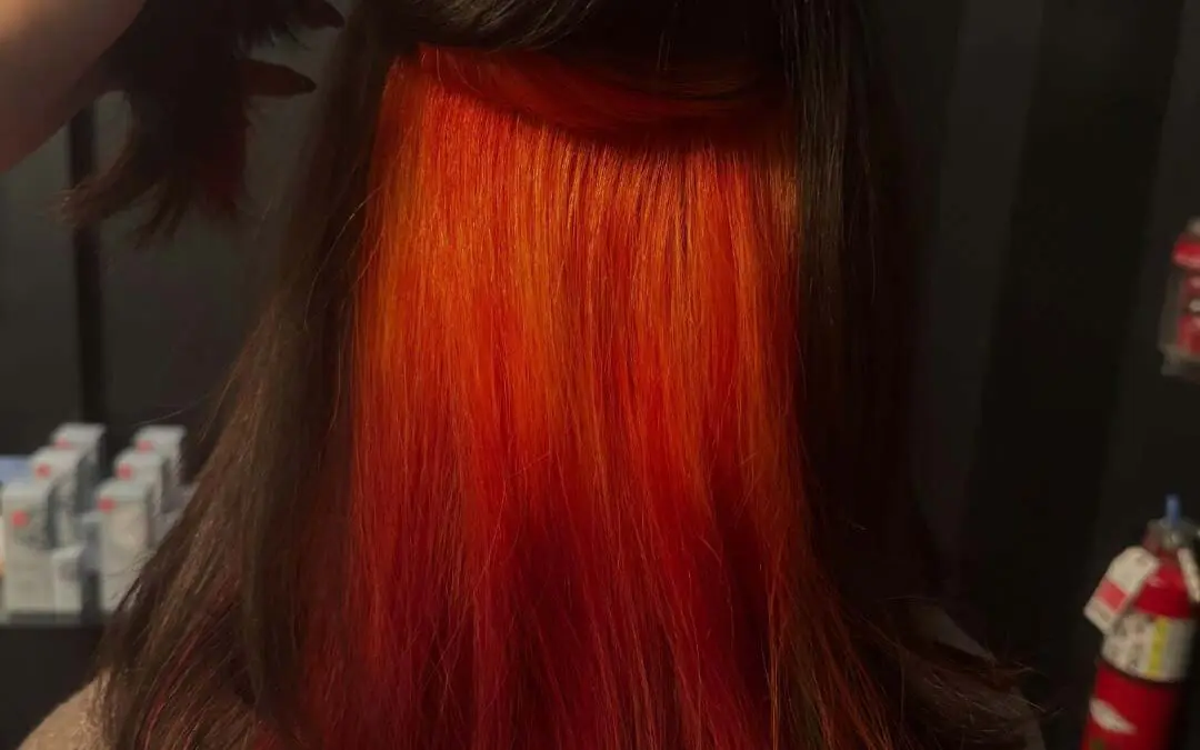 A client holding up a sleek dark brown top layer of shoulder-length hair to reveal a bold, vibrant fiery orange and red peekaboo underlayer, expertly crafted at Sunday Salon in Raleigh.