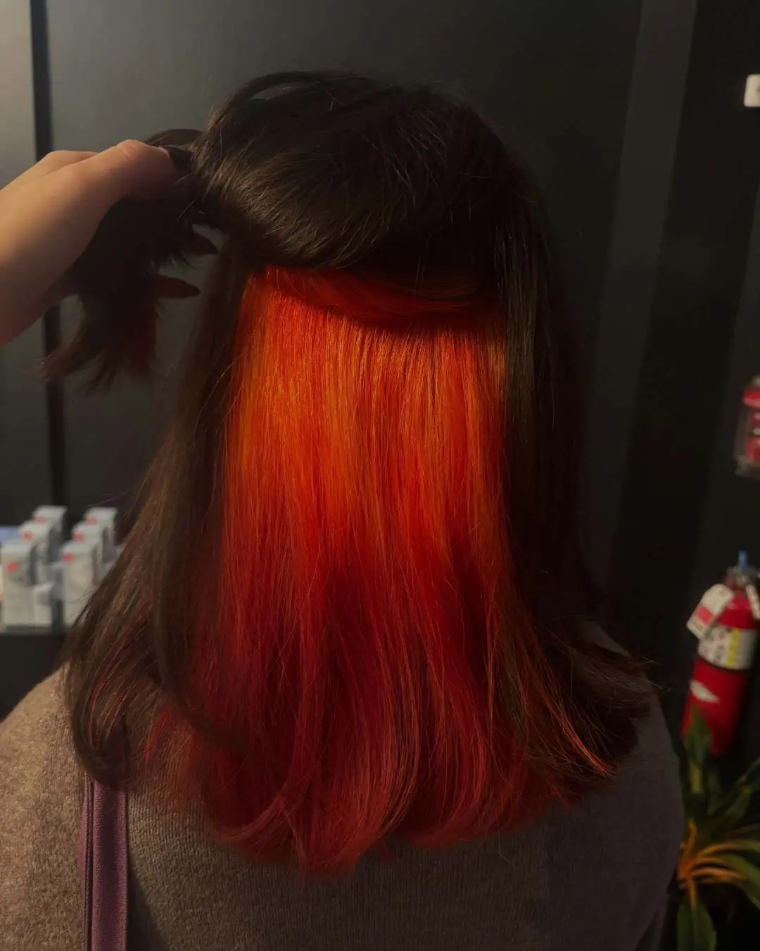 Fiery Orange Vivid Underlights with Dark Brunette Base A client holding up a sleek dark brown top layer of shoulder-length hair to reveal a bold, vibrant fiery orange and red peekaboo underlayer, expertly crafted at Sunday Salon in Raleigh.