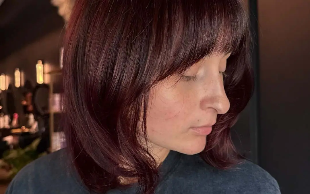 A client with a dark burgundy shoulder-length lob haircut featuring full face-framing bangs, soft texture, and rich mahogany tones, beautifully styled at Sunday Salon in Raleigh.