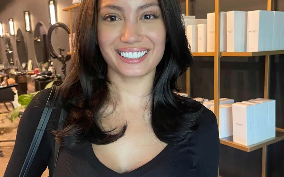 A smiling client at Sunday Salon in Raleigh showcasing mid-length, deep espresso brunette hair with a remarkably glossy finish. The hair is styled in a sleek, voluminous blowout featuring soft, bouncy face-framing layers that enhance its natural thickness and radiant shine.