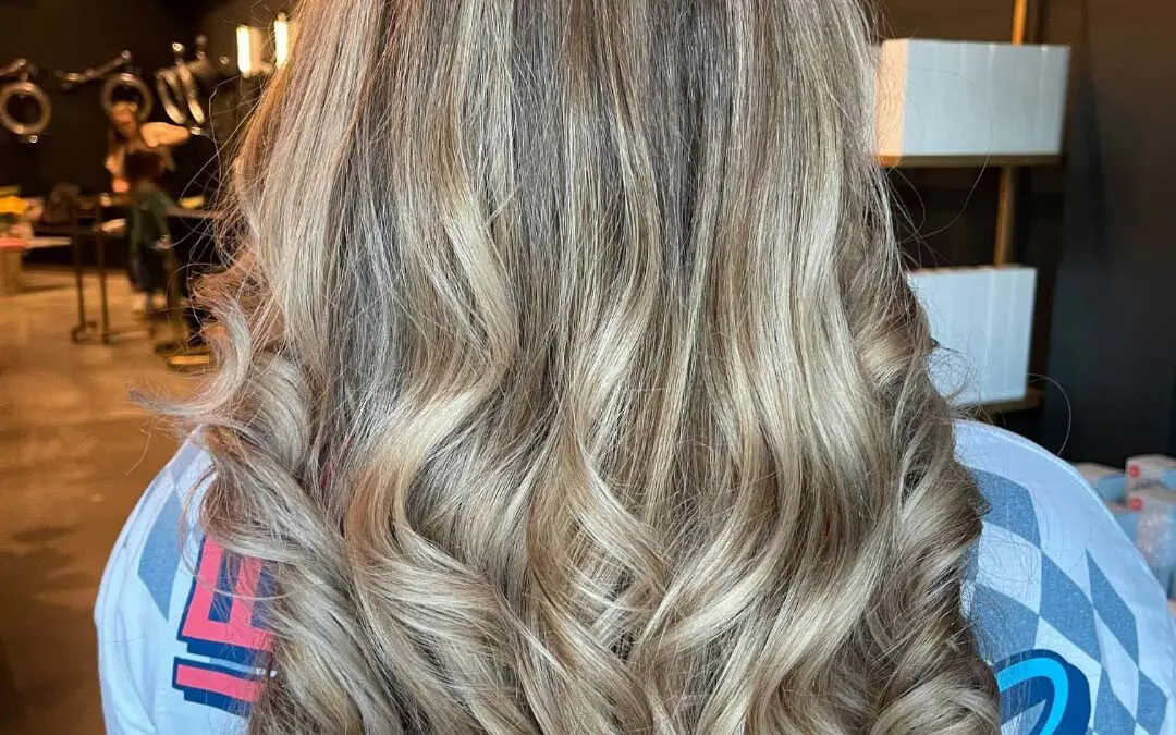 The back of a client's head at Sunday Salon in Raleigh, showcasing long, thick hair with multidimensional cool ash blonde highlights. The hair is styled smooth at the crown and cascades into beautifully defined, bouncy curls at the ends, highlighting the seamless blend and high-shine finish.