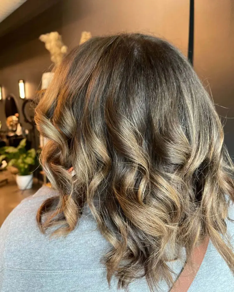 A beautifully styled client at Sunday Salon in Raleigh showcasing mid-length hair with soft, loose curls. The rich dark brunette base is seamlessly elevated with warm caramel and honey highlights, creating stunning dimension and a glossy, healthy finish that catches the light perfectly.