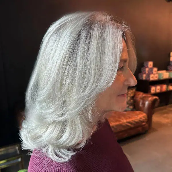grey-2 A woman with shoulder-length, wavy gray hair is shown in profile, wearing a maroon top. She is indoors, with a brown leather sofa and shelves visible in the dimly lit background.