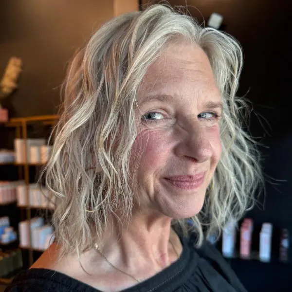 grey-5 A woman with wavy, shoulder-length gray hair and light skin smiles slightly, wearing a black top. Shelves with various products are blurred in the background. The lighting is soft and warm.