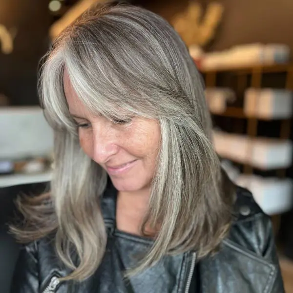 grey-6 A woman with long, straight gray hair and side-swept bangs looks down and smiles gently. She is wearing a black leather jacket, and the background appears to be a modern indoor setting.