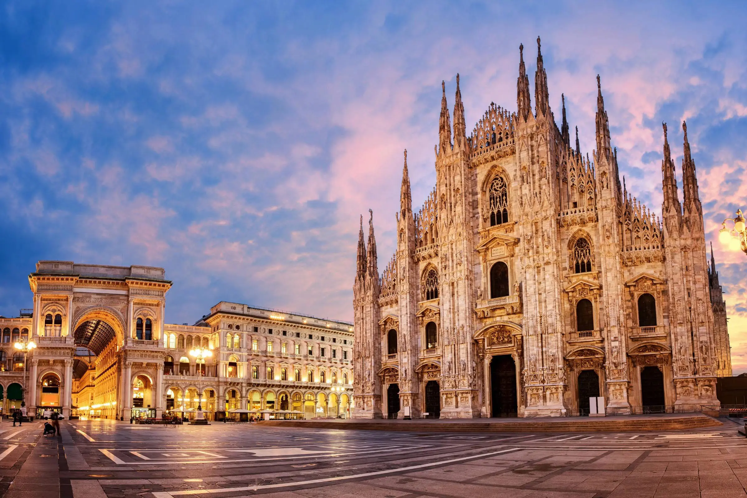 The image shows Milan Cathedral (Duomo di Milano) at sunset, its ornate Gothic façade and spires offering a stylist experience. The nearby Galleria Vittorio Emanuele II and surrounding buildings glow under warm streetlights in an empty square.